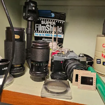 Vintage Minolta XG-7 SLR camera with various lenses, a Vivitar tripod head, a Cokin filter, and other photography accessories on a shelf.