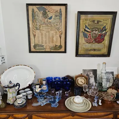 Assorted vintage collectibles displayed on a wooden sideboard, including World War I framed rolls of honor, cobalt blue glassware, a...