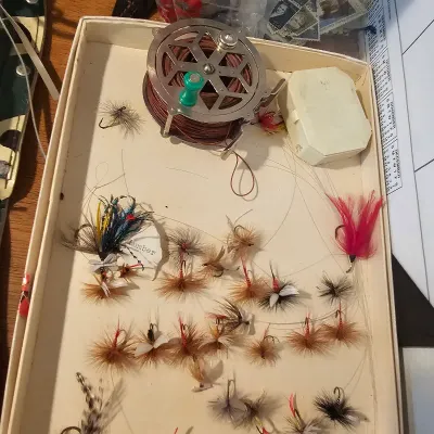 Vintage fly fishing collection featuring a metal fly reel with line, a small plastic fly box, and an assortment of hand-tied fishing...