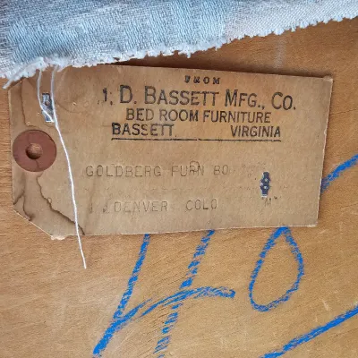 Vintage J.D. Bassett Mfg. Co. paper shipping tag attached to the wooden back of a bedroom furniture piece, showing delivery to Goldberg...