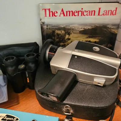 Vintage Super 8 movie camera resting on its hard case, displayed alongside Nikon Travelite II binoculars and "The American...