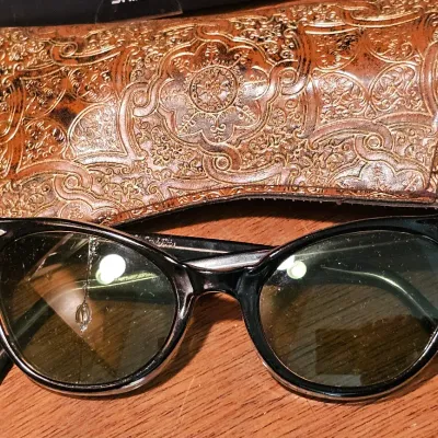 Vintage mid-century cat-eye sunglasses with black frames and green-tinted lenses, shown with an embossed brown leather King Optical Co....