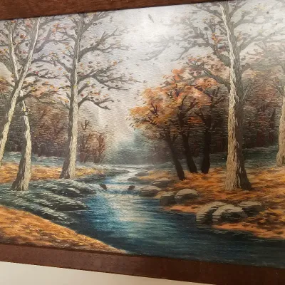 Framed vintage embroidery art depicting a serene autumn forest landscape with a flowing stream, housed in a simple dark wood frame.