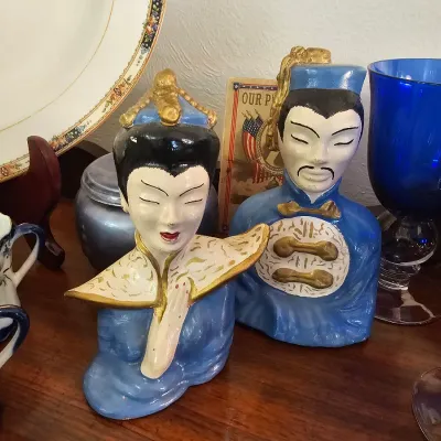 Pair of mid-century ceramic Asian bust figurines in blue and gold attire, displayed alongside vintage glassware and teacups.
