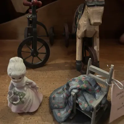 Sale Photo Thumbnail #43: Assorted vintage-style decorative figurines, including a porcelain girl holding flowers, a miniature rocking chair with a quilt, a metal tricycle, and a carved wooden horse tricycle.