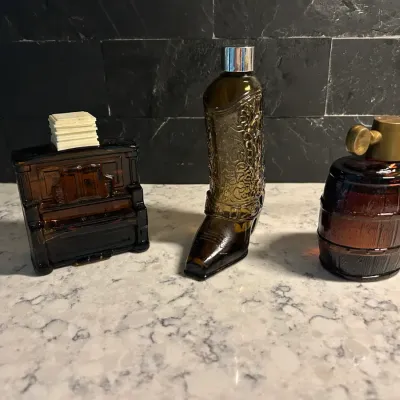 Sale Photo Thumbnail #116: Three vintage Avon amber glass cologne bottles, featuring a piano, a cowboy boot, and a barrel shape, circa 1970s.