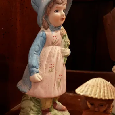 Sale Photo Thumbnail #36: Vintage hand-painted bisque porcelain figurine of a young girl in a blue bonnet and pink apron holding flowers, showing some dust from storage.