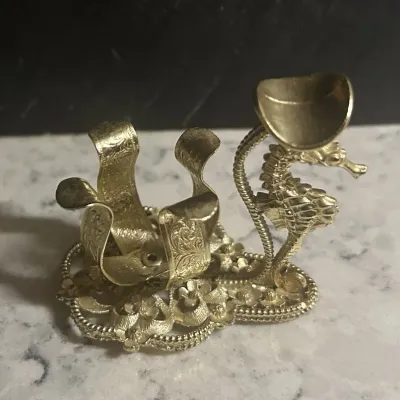 Sale Photo Thumbnail #154: Vintage gold-tone metal lipstick holder featuring an ornate filigree base and a decorative seahorse figure.