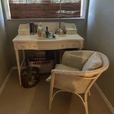 Sale Photo Thumbnail #115: White painted wooden vanity desk with matching wicker armchair, featuring a small desk lamp and various perfume bottles on top.