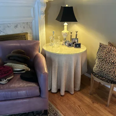 Mauve leather armchair holding a collection of hats, paired with a skirted round side table displaying a lamp and picture frames, next to...