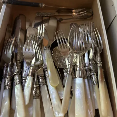 Assorted vintage mother-of-pearl handled flatware, including forks and knives with ornate silver-plated ferrules, stored in a cardboard box.