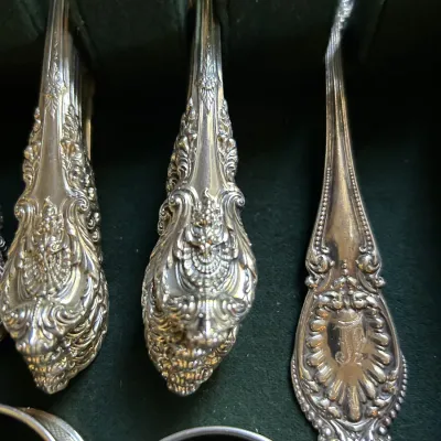Ornate sterling silver flatware handles featuring intricate scrollwork and floral motifs, stored in a green felt-lined chest.