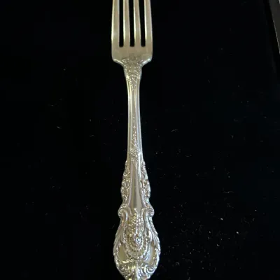 Sterling silver dinner fork featuring an ornate, vintage floral pattern on the handle, displayed against a black background.