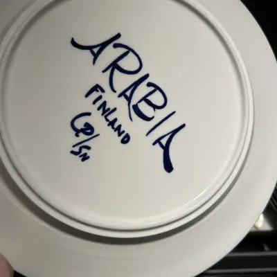 Hand-painted maker's mark on the underside of an Arabia Finland ceramic plate, featuring the artist's initials.