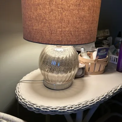Sale Photo Thumbnail #114: Contemporary table lamp featuring a ribbed mercury glass base and a brown fabric drum shade, displayed on a white wicker table.