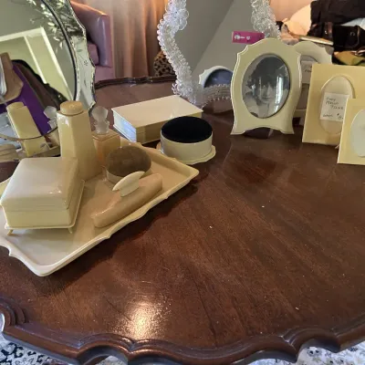 Vintage celluloid vanity set, including a tray, lidded boxes, a brush, and picture frames, displayed on a scalloped mahogany table.