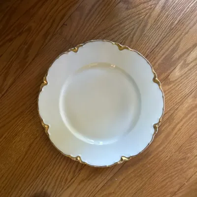 Vintage white porcelain plate featuring a scalloped rim with elegant gold trim, shown on a hardwood floor.