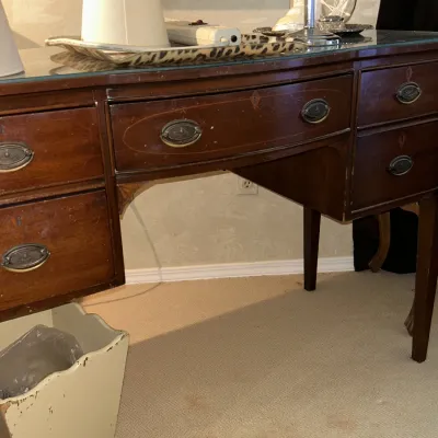 Sale Photo Thumbnail #113: Vintage mahogany vanity desk featuring a curved front, glass top protector, and brass bail pulls, showing some surface wear.
