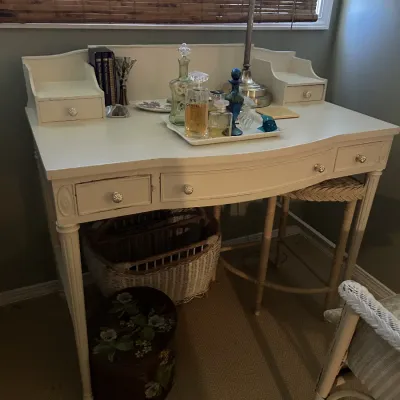 Sale Photo Thumbnail #116: Vintage white-painted wooden vanity desk with a curved front, fluted legs, and matching upper organizer drawers, styled with perfume bottles and a table lamp.