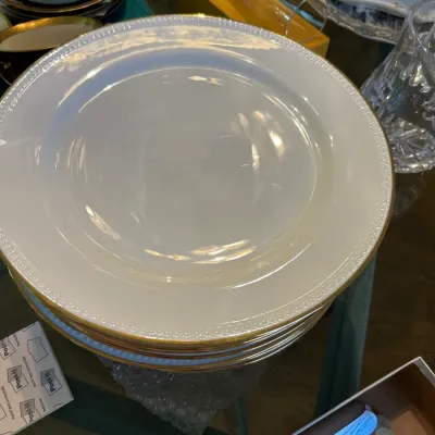 Stack of white porcelain dinner plates featuring a delicate textured rim and gold trim, shown alongside other glassware and tableware.