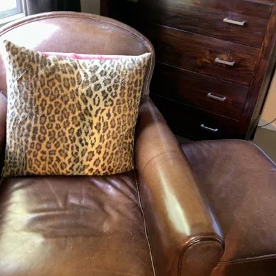 Sale Photo Thumbnail #111: Vintage brown leather armchair and matching ottoman, paired with a leopard print throw pillow, positioned next to a dark wood four-drawer chest.