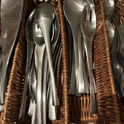 Mid-century modern stainless steel flatware set, including spoons, forks, and serving utensils, organized in a woven wicker basket.