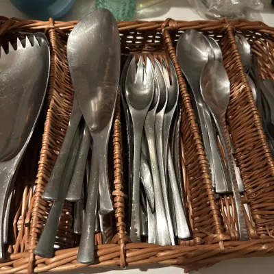 Mid-century modern stainless steel flatware set, including serving utensils and forks, organized in a woven wicker basket.