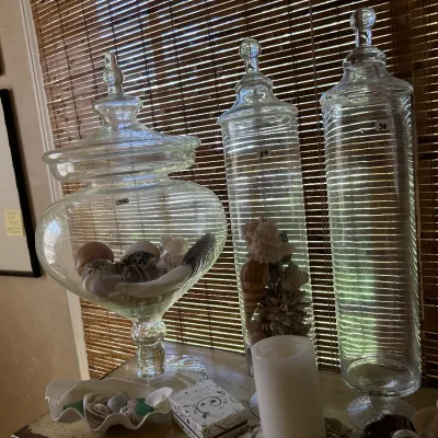 Three clear glass apothecary jars of varying heights and shapes, filled with decorative seashells and displayed on a tabletop.