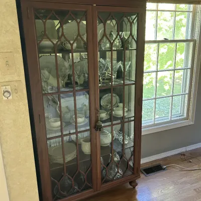 Sale Photo Thumbnail #123: Vintage mahogany china cabinet featuring a broken pediment top with a central finial, glass doors with gothic-style mullions, and bun feet, filled with glassware and china.