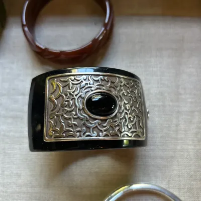 Vintage black acrylic cuff bracelet featuring an ornate silver-tone filigree plaque set with a central black cabochon, shown alongside...