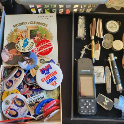 Sale Photo Thumbnail #205: Collection of vintage political campaign buttons, including Clinton/Gore '92, alongside a tray of pocket knives, a compass, a Lowrance GPS unit, and a leather-sheathed hunting knife.