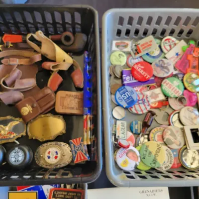 Sale Photo Thumbnail #204: vintage collectibles, featuring belt buckles, and leather holsters on the left, and an assortment of pinback buttons, keychains, and small novelty items on the right.