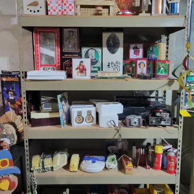 Sale Photo Thumbnail #153: Five-tier metal shelving unit filled with various collectibles, including Elvis Presley memorabilia, vintage cameras, electronics, and holiday decorations.