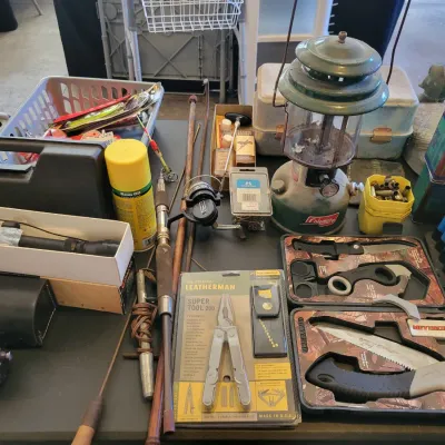 Sale Photo Thumbnail #187: Assortment of outdoor and hunting gear, including a vintage Coleman lantern, a Leatherman Super Tool 200, various knives, fishing reels, and shells.