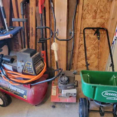 Sale Photo Thumbnail #196: Coleman Powermate 11-gallon air compressor, a small gas-powered tiller, and a green Scotts SpeedyGreen 1000 lawn spreader.