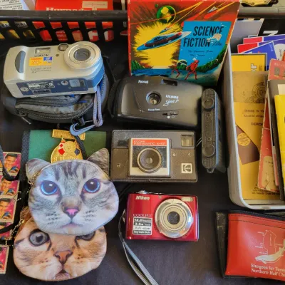 Sale Photo Thumbnail #206: Assortment of vintage and digital cameras, including a red Nikon Coolpix, a silver Sony Cyber-shot, a Kodak Instamatic X-15F, and a Kalimar Spirit P, alongside cat-face coin purses and various ephemera.