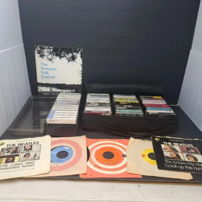 Collection of vintage music media, including cassette tapes in a black carrying case, a Newport Folk Festival program, and several 45 RPM...