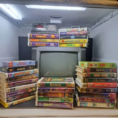 Vintage CRT television surrounded by stacks of VHS tapes, featuring classic Disney and family films from the 1990s and early 2000s.