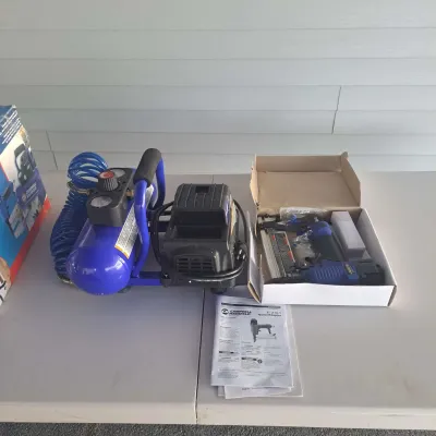 Campbell Hausfeld portable air compressor with coiled blue hose, accompanied by a 2-in-1 nailer/stapler in its original box with...