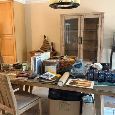 Rustic wooden dining table with a glass top and matching chairs, shown holding various estate sale items including a Moen faucet box,...