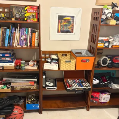 Three-piece wooden shelving unit holding various items including books, board games, electronics, and storage bins.