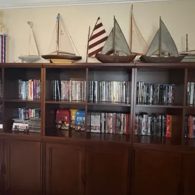 Large dark wood media console and shelving unit filled with DVDs, Blu-rays, and box sets, topped with a collection of model sailboats and...