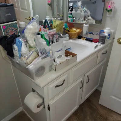 Sale Photo Thumbnail #19: White bathroom vanity cabinet with a molded sink top, featuring two doors, two drawers, and a side-mounted toilet paper holder, shown cluttered with various personal care items.