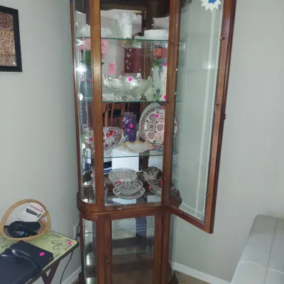 Sale Photo Thumbnail #4: Traditional wooden curio cabinet with curved glass sides, glass shelves, and interior lighting, filled with various glassware and decorative plates.
