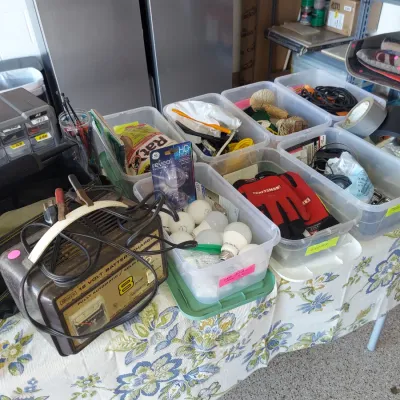 Sale Photo Thumbnail #11: Vintage Sears 12-volt battery charger alongside various household and garage items, including lightbulbs, Craftsman work gloves, and tape, organized in plastic bins.