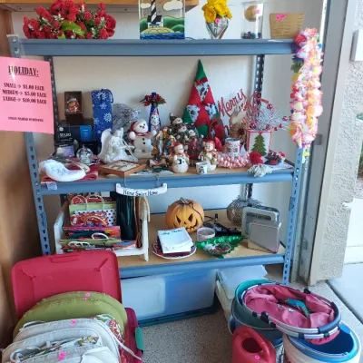 Sale Photo Thumbnail #24: Assorted holiday decorations and household items displayed on a metal shelving unit, including Christmas figurines, a Halloween pumpkin, gift bags, and outdoor cushions.