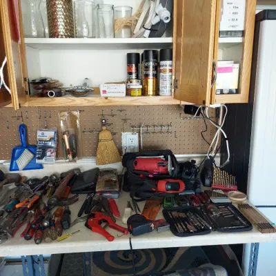 Sale Photo Thumbnail #12: Assorted hand tools, power tools, and workshop supplies arranged on a workbench with overhead cabinets and a pegboard organizer.
