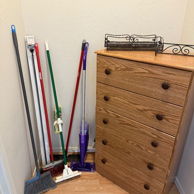 Oak-finish five-drawer dresser alongside an assortment of household cleaning supplies, including brooms and mops.
