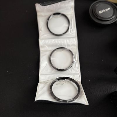 Sale Photo Thumbnail #4: Set of three vintage camera lens filters housed in a white vinyl folding pouch, shown alongside a Nikon lens cap.