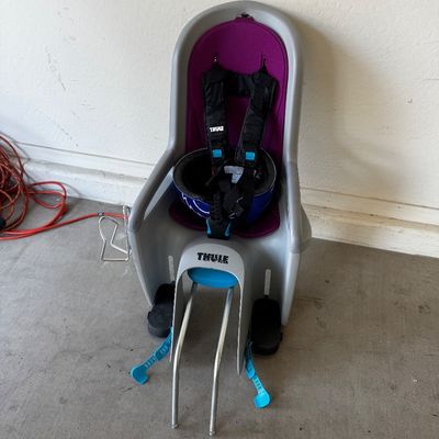 Sale Photo Thumbnail #78: Thule RideAlong child bike seat in gray and purple, featuring adjustable harness straps and footrests, shown with a blue helmet resting inside.